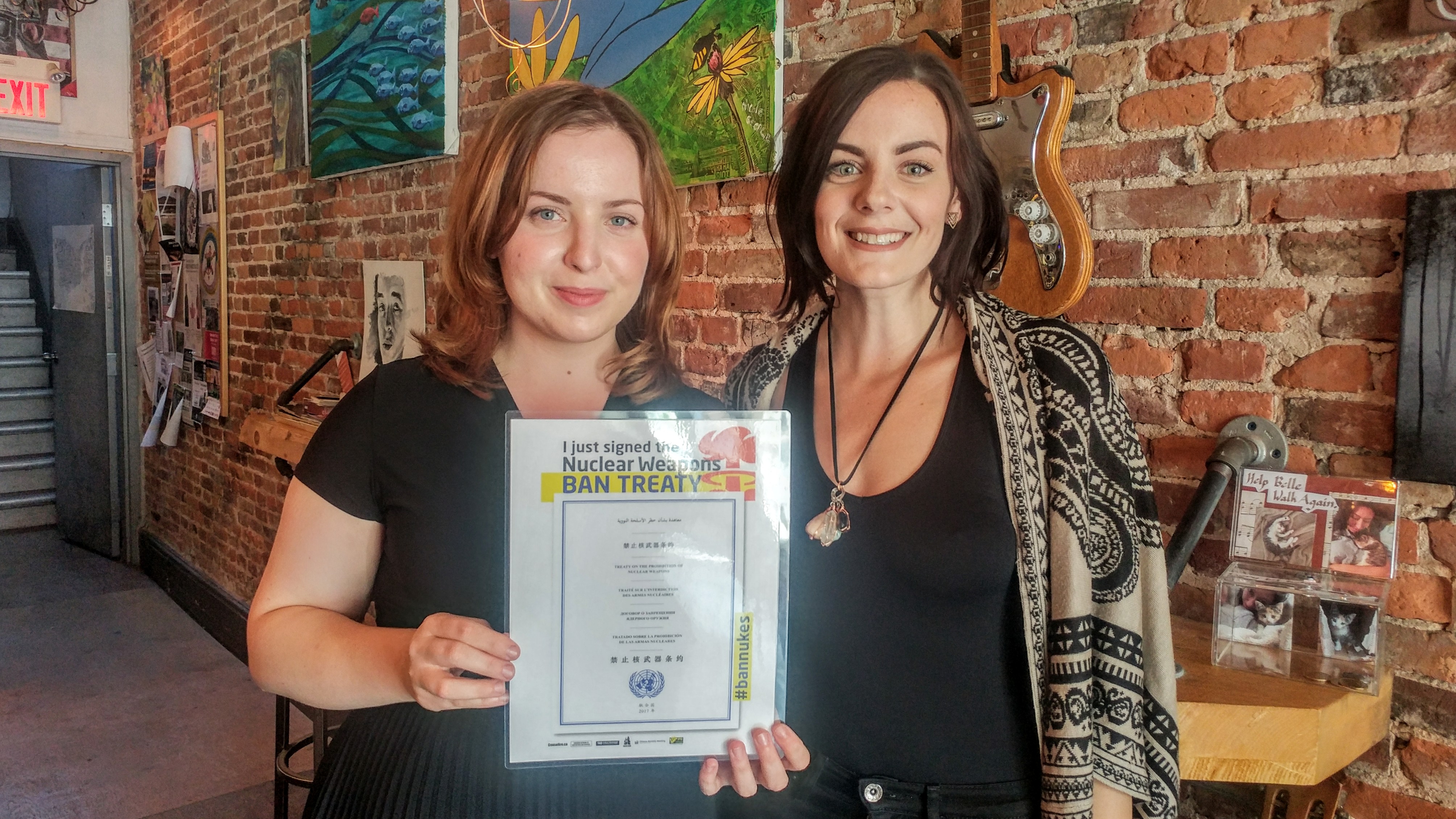 Two young women in a cafe hold up a sign which reads in several languages "I just signed the Nuclear Weapons Ban Treaty"