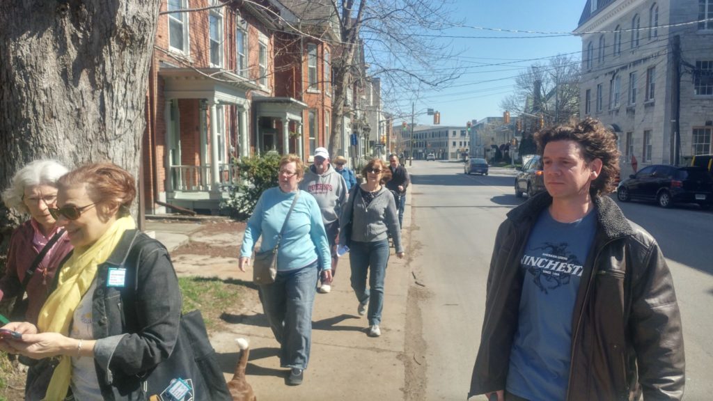 Three people in the foreground seen waist-up and six people behind on the sidewalk on a stretch of Bagot St between stops 4 and 4A.