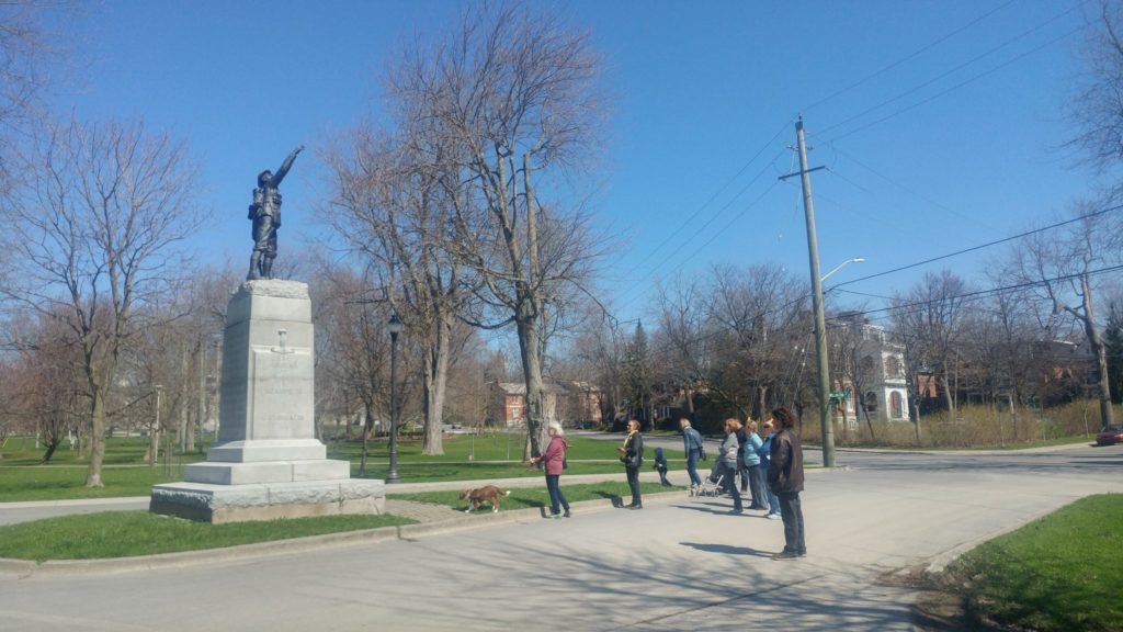 Eight individuals and a dog stand dwarfed before the twenty foot tall WWI infantry memorial in City Park, Stop 5 on the tour. 